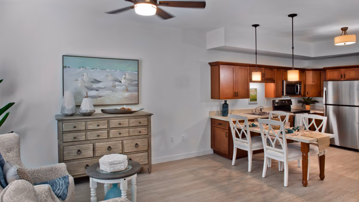 Open-plan interior with a living area (armchair and dresser with seagull painting) adjacent to a kitchen and dining table with wooden cabinets and stainless steel appliances.