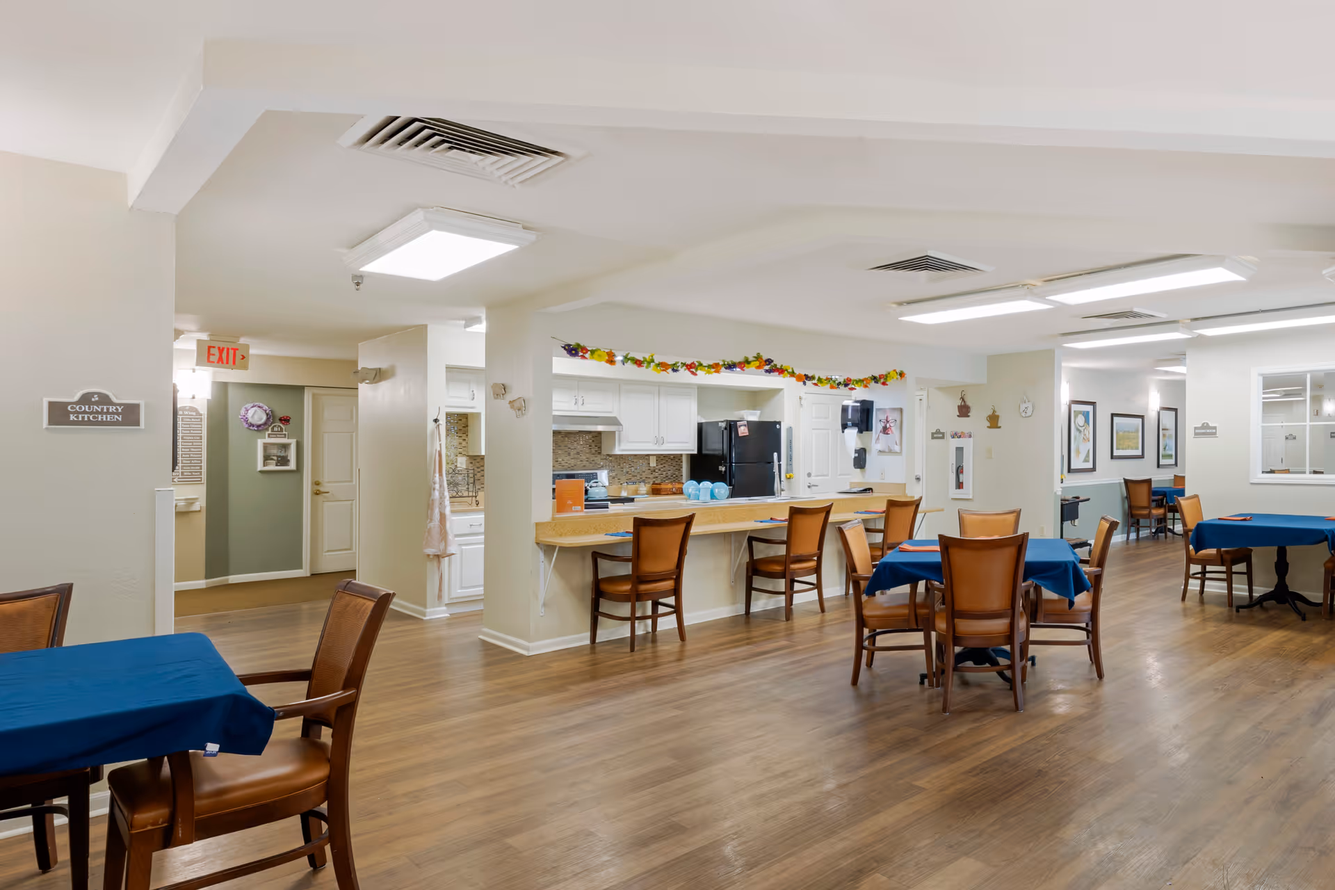 Interior view of a senior living facility dining area with wooden floors, tables covered with blue tablecloths, and wooden chairs. There is a kitchen area with a counter and bar stools, white cabinets, a black refrigerator, and a stove. The walls are decorated with framed pictures and a garland of flowers above the kitchen counter. An exit sign and a sign reading 'Country Kitchen' are visible on the left wall.