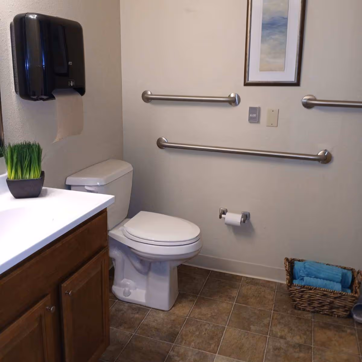 Accessible bathroom showing a toilet, sink vanity, wall-mounted grab bars, a paper towel dispenser, and a basket of towels on a tiled floor.