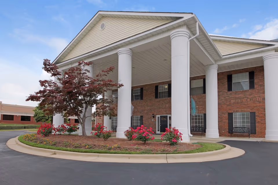 Front exterior view of a senior living facility with large white columns supporting a covered entrance. The building has red brick walls, black window shutters, and a landscaped area with a tree and red flowers in front. The sky is partly cloudy.