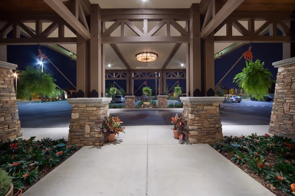 Night view of the entrance area of a facility with stone pillars supporting a wooden roof structure. Hanging green plants are suspended from the beams, and potted plants are placed near the pillars. The area is well-lit with a ceiling light and outdoor lighting, with a parking lot and cars visible in the background.