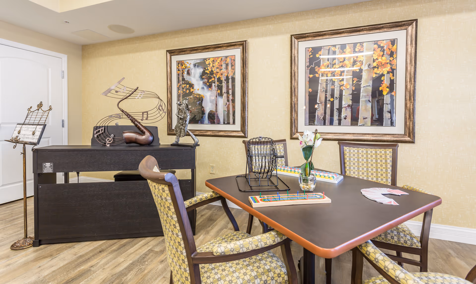 A cozy activity room with a table and four patterned chairs. On the table are a bingo cage, bingo cards, a vase with flowers, and a cribbage board. Behind the table is a piano with a music stand and decorative musical notes sculpture. The walls are light yellow with two framed paintings of autumn trees.