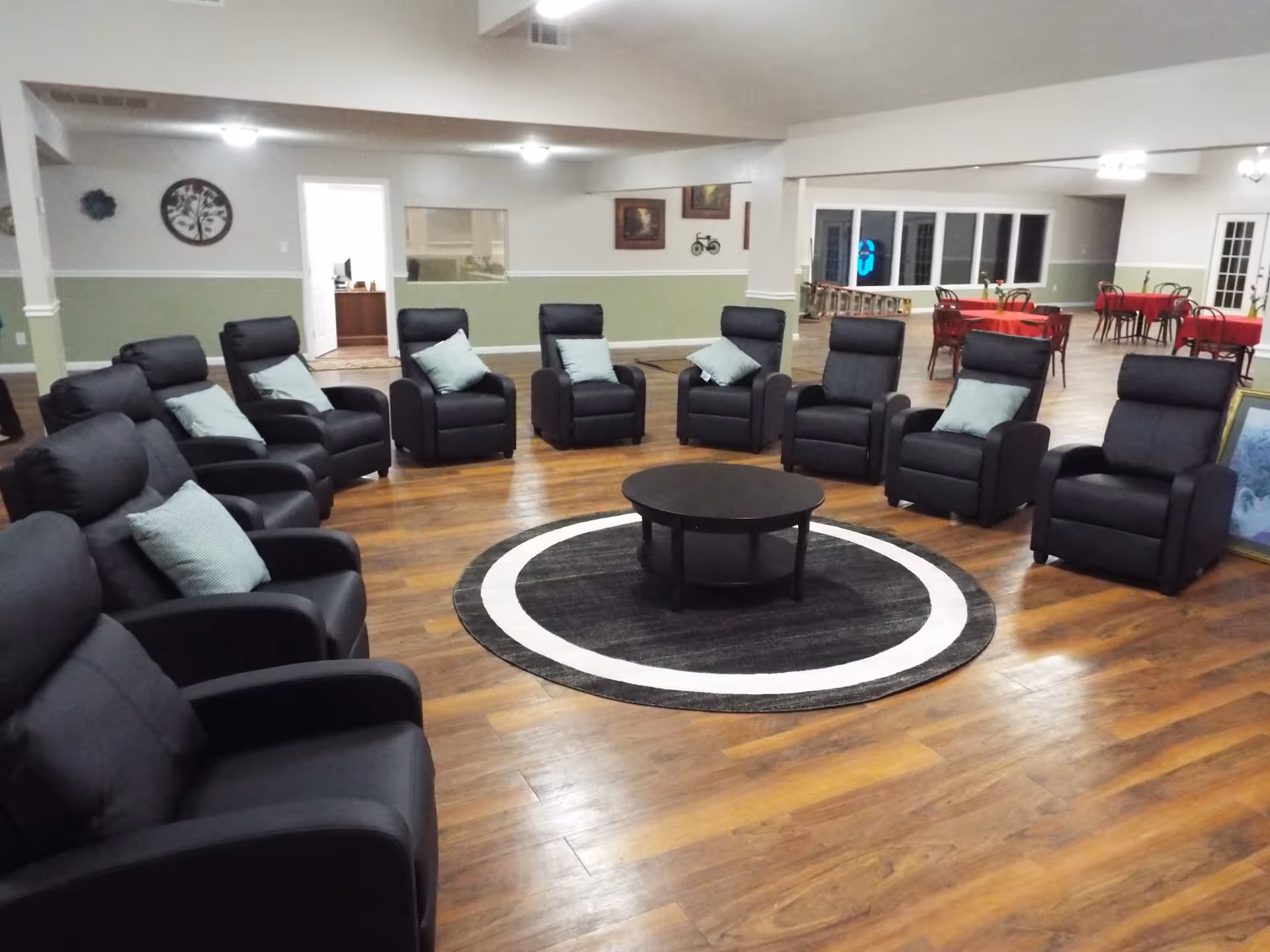 A spacious common area with a circle of black recliner chairs each with a light blue cushion, surrounding a round black coffee table on a circular black and white rug. The room has wooden flooring, light green and white walls, and several tables with red tablecloths in the background. There are framed pictures on the walls and a door leading to another room.