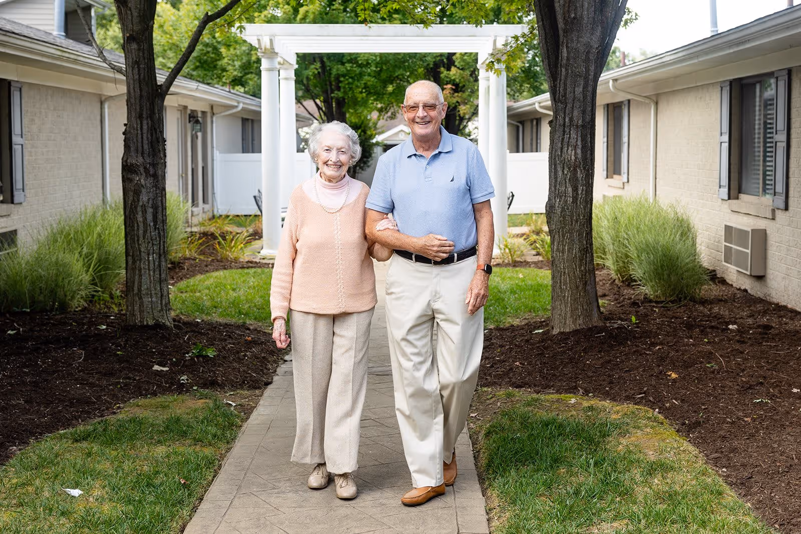 An elderly couple walking arm in arm on a paved pathway between two buildings with trees and greenery around them. The woman is wearing a peach sweater and beige pants, and the man is wearing a light blue polo shirt and beige pants. Both are smiling and appear happy.