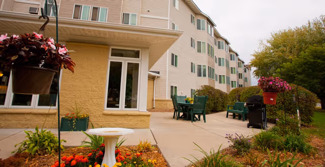 Outdoor patio area at Blaine Courts with green chairs and tables, a barbecue grill, hanging flower pots, and landscaped garden beds with colorful flowers in front of a multi-story beige building with green window shutters.
