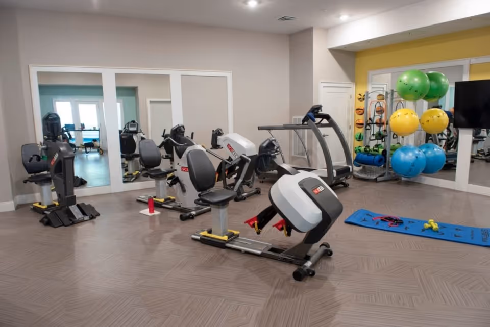 A fitness room with exercise equipment including recumbent bikes, a treadmill, a rack of colorful exercise balls, yoga mats, and small weights. The room has large mirrors on the walls and a television mounted on the right side.