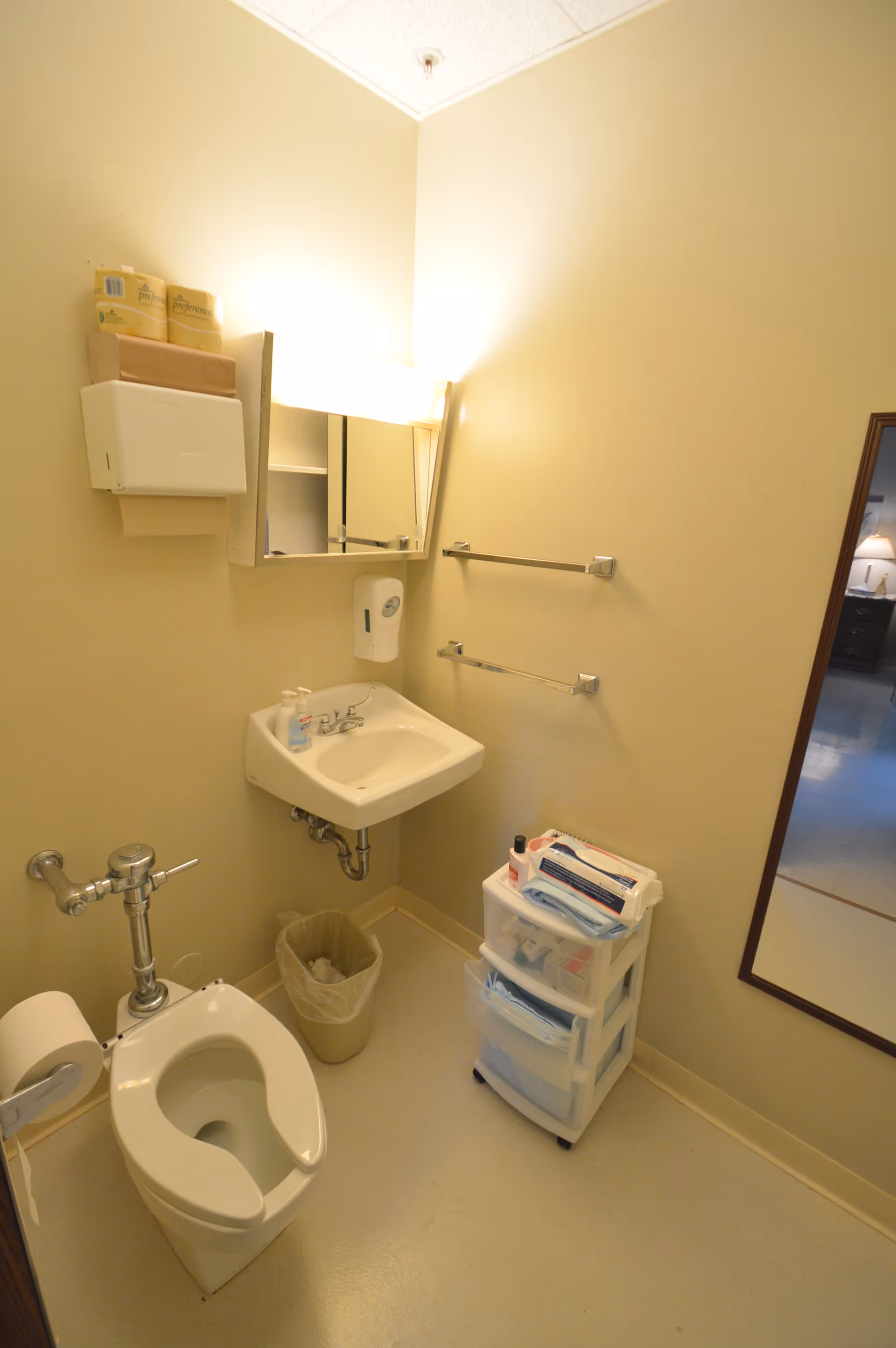 Small beige institutional bathroom with a toilet, wall-mounted sink and mirror, towel racks, and a plastic storage drawer cart.