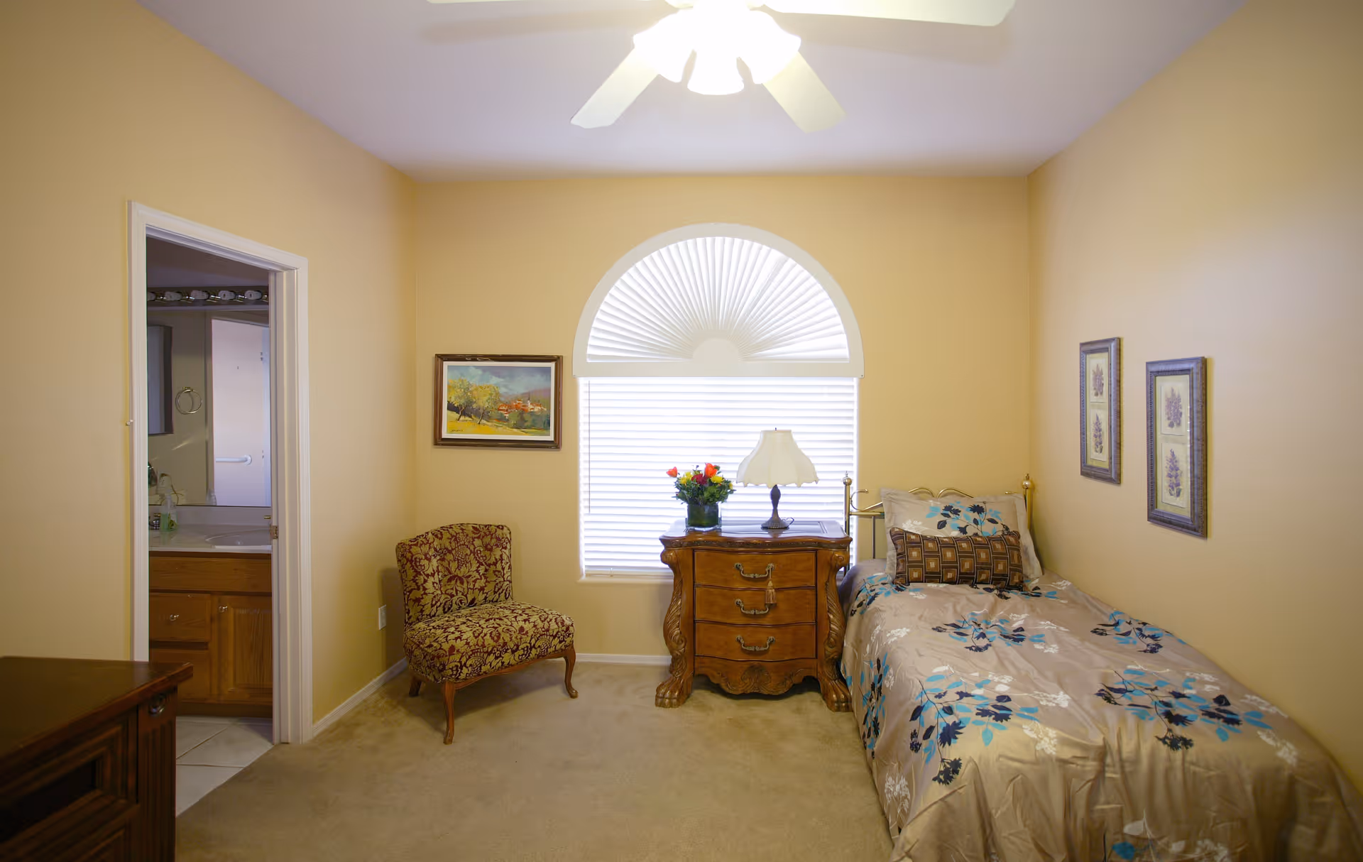 A small bedroom with beige walls and carpet. There is a single bed with floral bedding on the right side, a wooden nightstand with a lamp and a vase of flowers in front of a window with a half-circle fan-shaped blind. A patterned upholstered chair is placed near the left wall, and a doorway leads to a bathroom with a sink and mirror visible.
