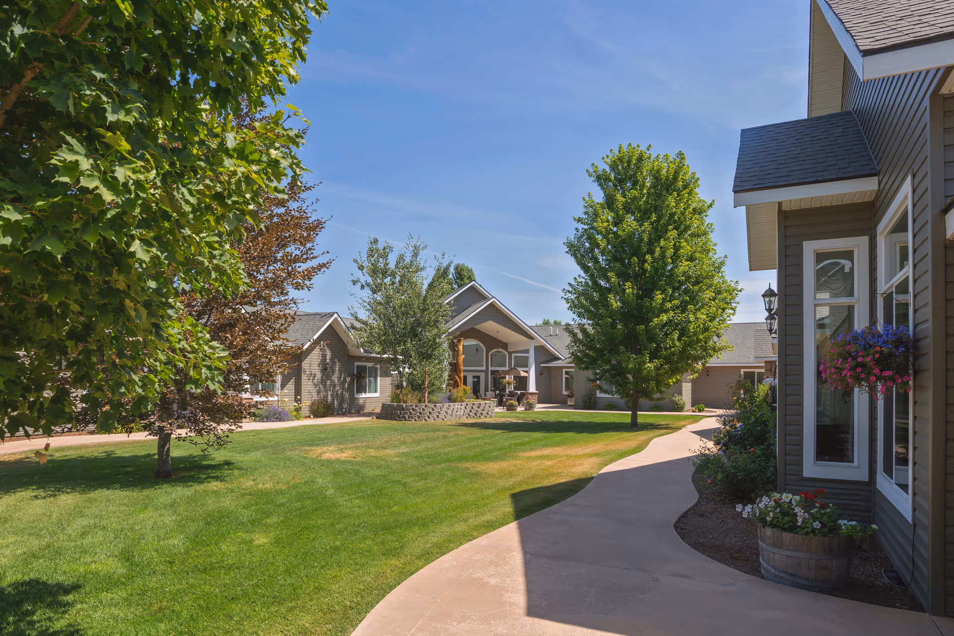 A sunny outdoor view of a senior living facility with a curved concrete walkway, green grass, several trees, and buildings with gray siding and large windows. Flower pots with colorful flowers are placed near the building on the right.
