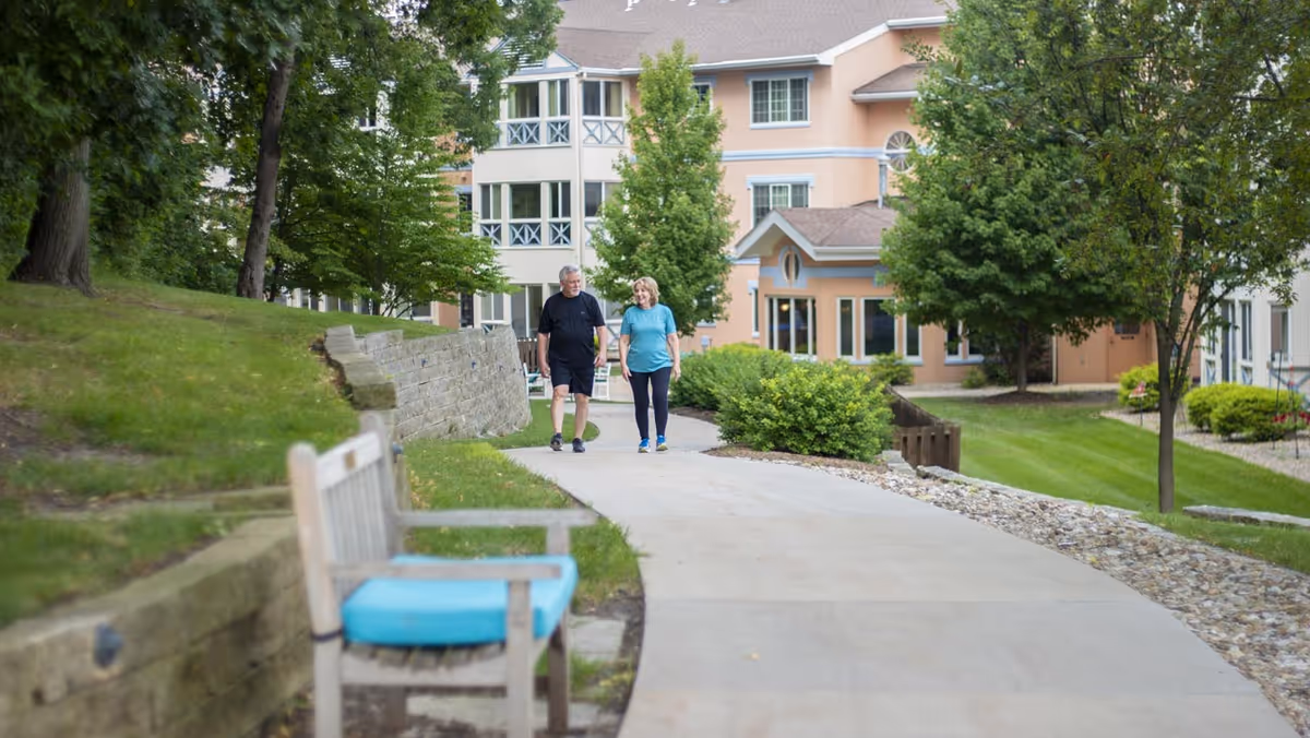 A man and a woman walking together on a paved pathway surrounded by green grass, trees, and bushes. There is a wooden bench with a blue cushion on the left side of the path. In the background, there is a multi-story building with beige and white exterior walls and several windows.