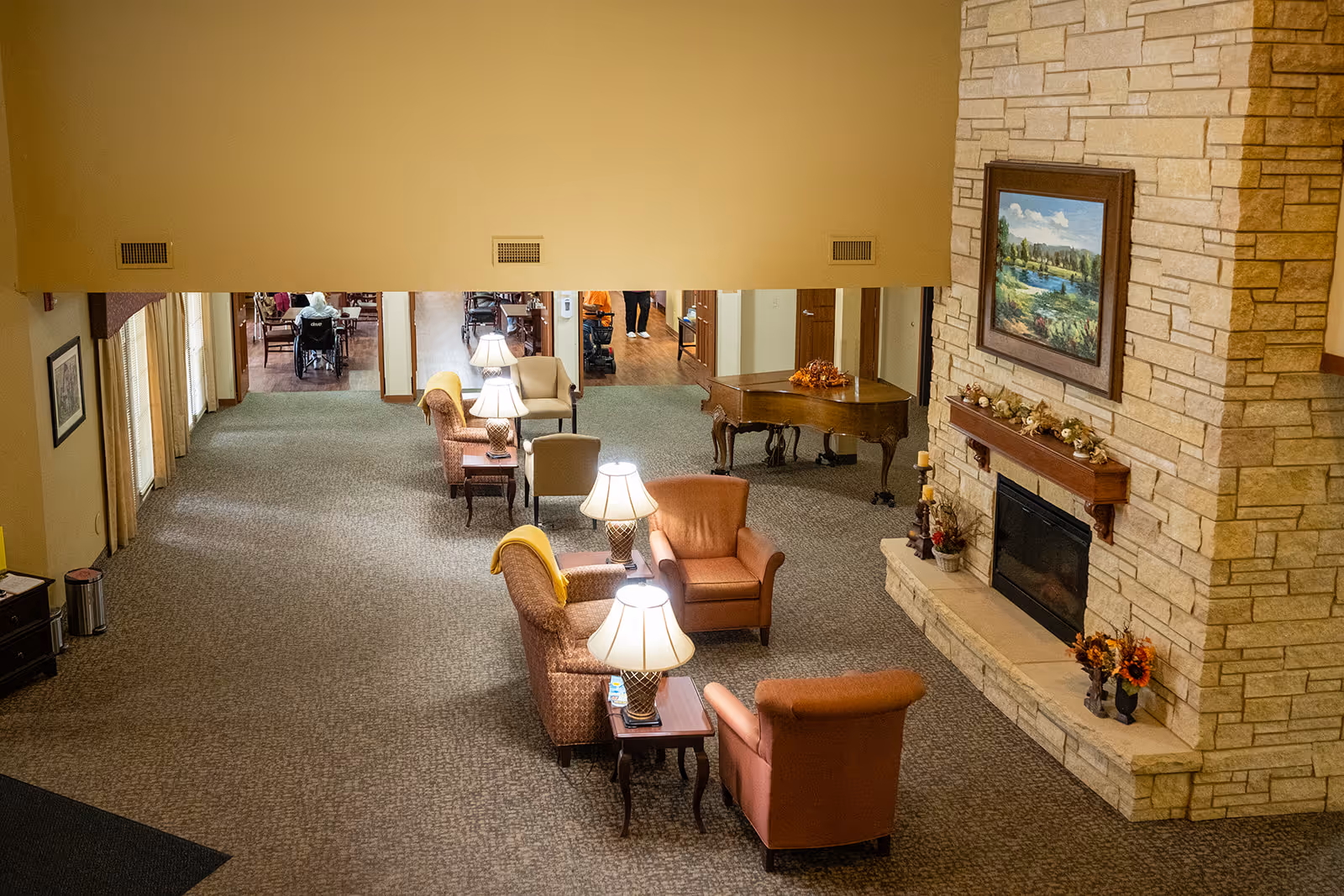 A spacious common area in a senior living facility featuring a stone fireplace with a framed landscape painting above it. The room has several armchairs and sofas arranged around small tables with lamps. In the background, there is a grand piano and a dining area with people seated at tables.