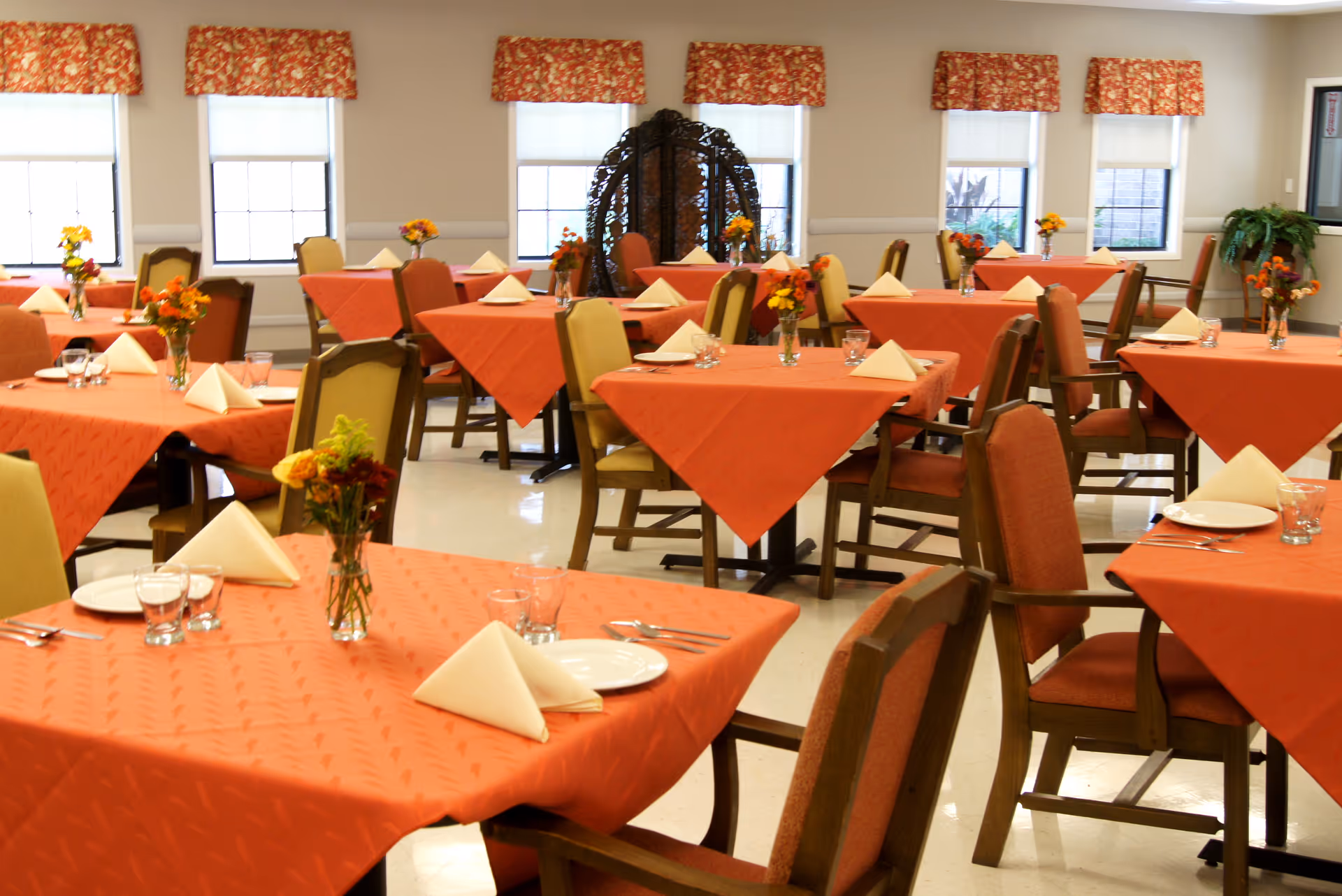Dining room with multiple tables covered in orange tablecloths, each set with white folded napkins, plates, glasses, and silverware. Small floral centerpieces are placed on each table. The room has several windows with patterned valances and cushioned chairs around the tables.