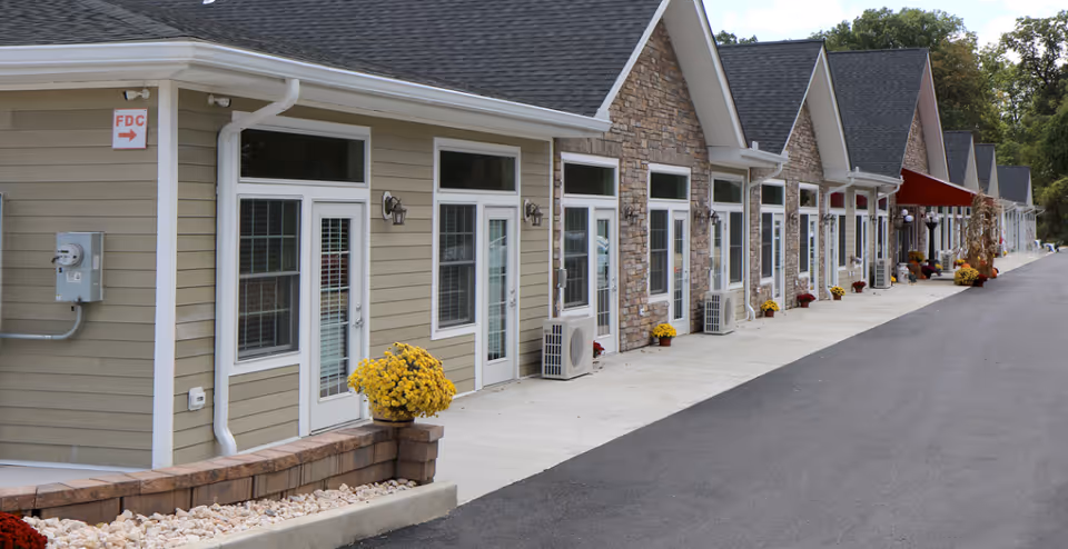 Row of beige cottage-style senior living units with glass doors lining a paved walkway.