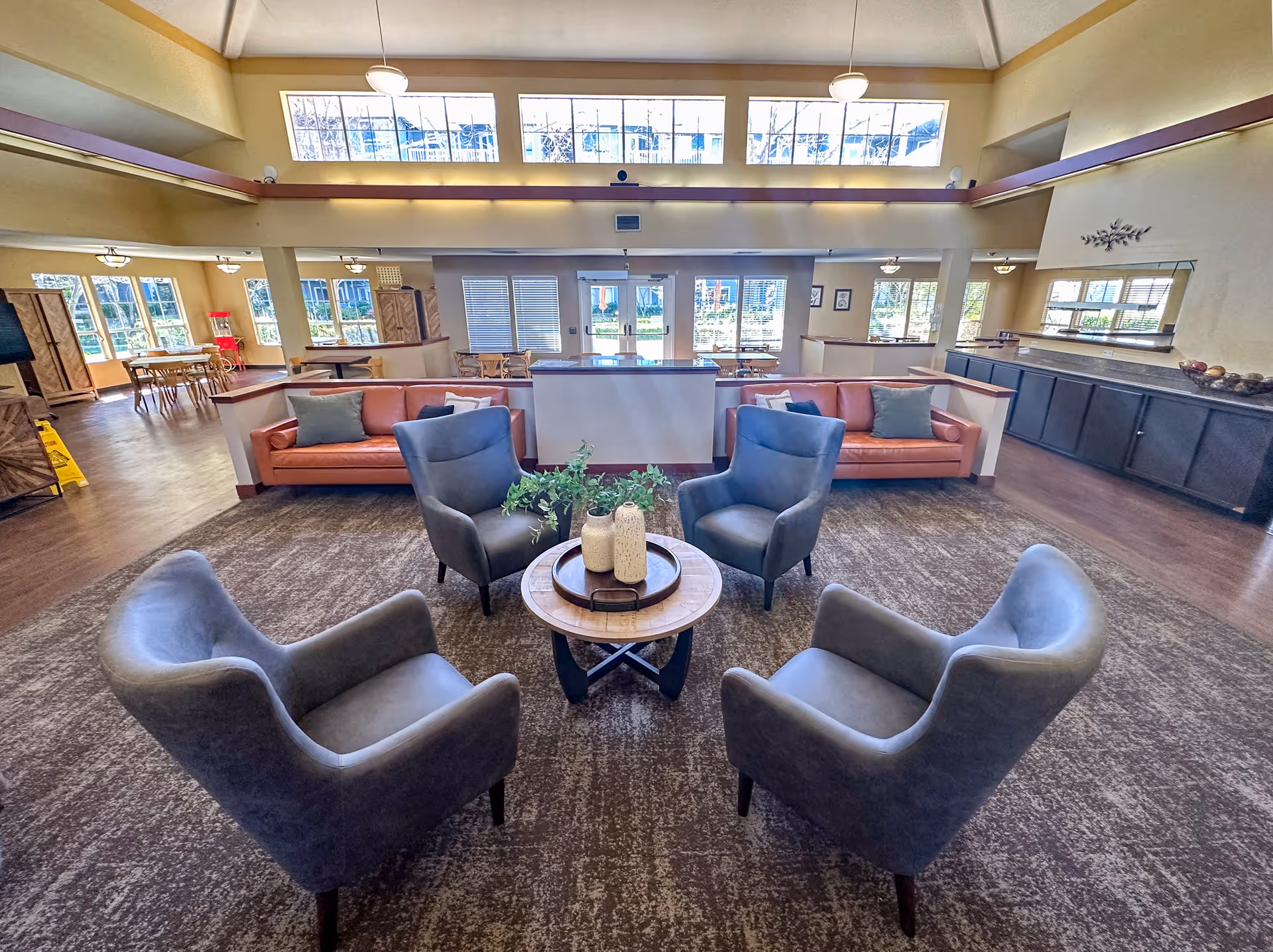 A spacious and well-lit common area with four gray armchairs arranged around a round wooden coffee table with decorative vases and greenery. Behind the chairs are two orange sofas with pillows, separated by low walls. Large windows and glass doors let in natural light, and there are additional tables and chairs in the background. The room has high ceilings with hanging lights and a carpeted floor area surrounded by wood flooring.