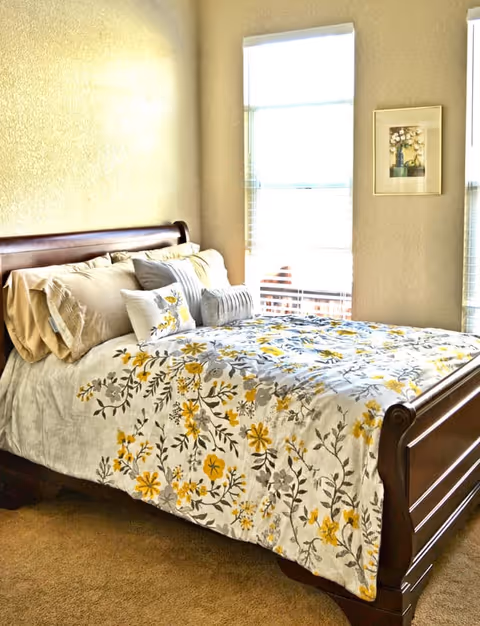 A cozy bedroom featuring a wooden sleigh bed with a floral patterned bedspread in yellow and gray tones, multiple pillows, beige walls, and two tall windows with blinds letting in natural light. A framed floral artwork hangs between the windows.
