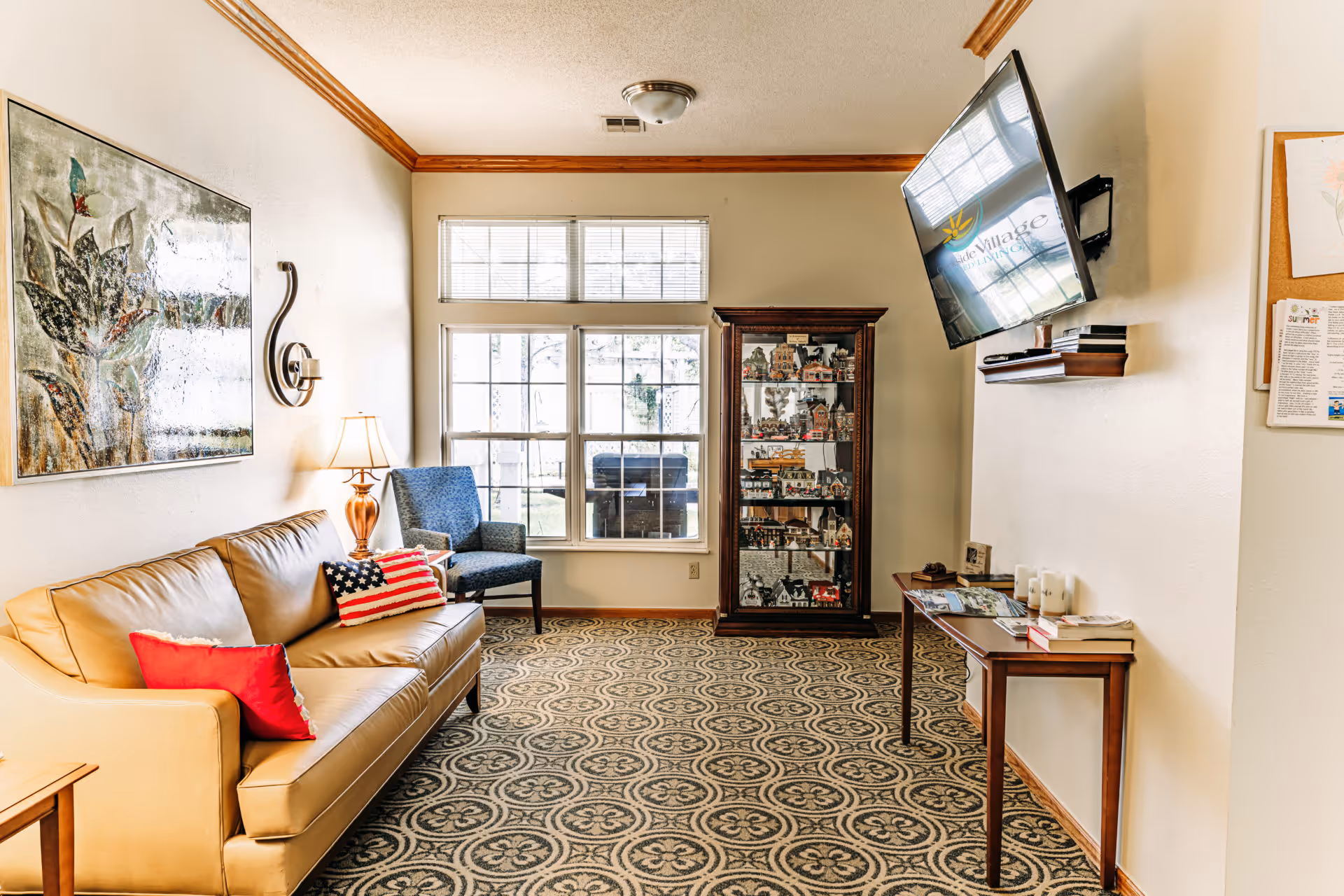 A cozy living room area with a tan leather sofa adorned with red and American flag pillows, a blue upholstered chair, a wooden side table with a lamp, a patterned carpet, a glass display cabinet filled with miniature houses, a wall-mounted TV displaying the Creekside Village logo, and a small wooden table with books and magazines.