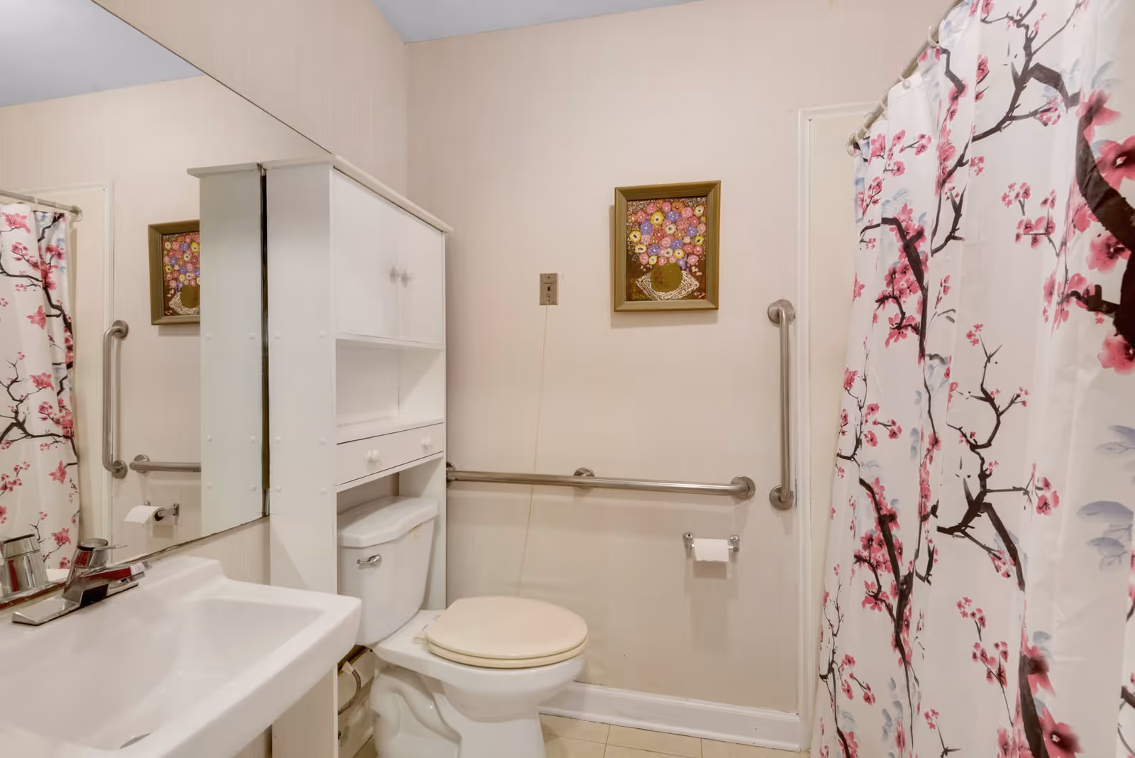 A bathroom with a white sink and faucet on the left, a white toilet with a beige seat, and a white cabinet above the toilet. There are metal grab bars on the walls near the toilet and shower area. The shower curtain has a floral design with pink and brown branches. A framed picture of colorful flowers hangs on the wall above the grab bar.
