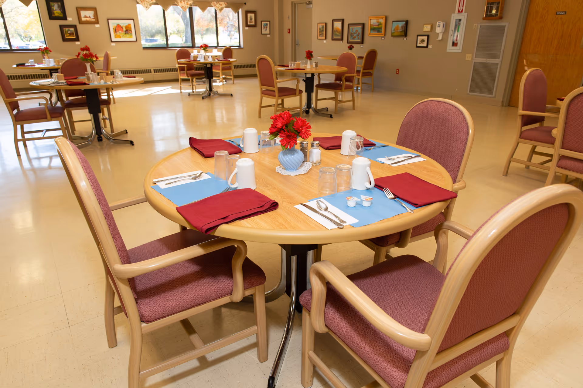 A dining room in a senior living facility with round wooden tables set for four people. Each table has place settings with utensils, glasses, mugs, red cloth napkins, and a small vase with red flowers. The room has large windows letting in natural light, framed artwork on the walls, and beige flooring.