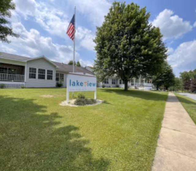 Exterior view of a single-story building with a large green lawn and a sidewalk. There is a sign in the grass that reads 'lakeview' with an American flag flying on a flagpole behind it. Trees line the sidewalk and the sky is partly cloudy.