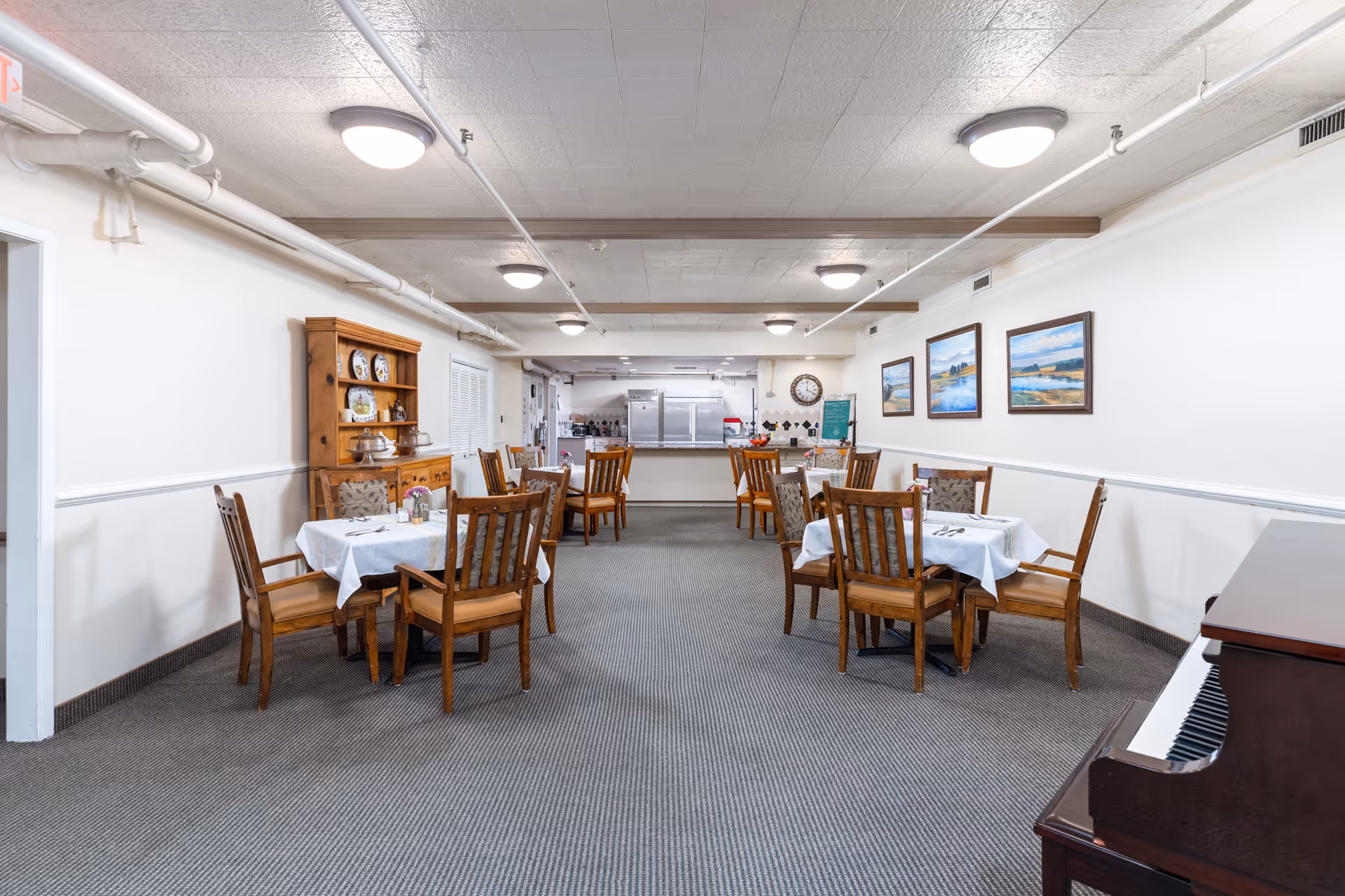 Dining room with multiple set tables and wooden chairs, a piano on the right, and a serving area/kitchen at the far end.