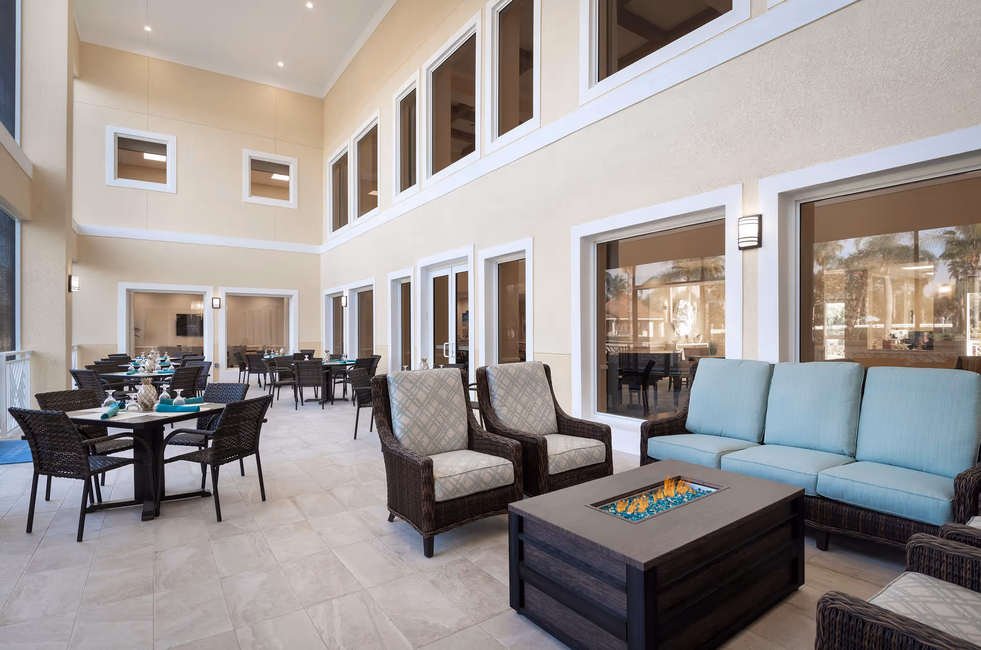 Indoor seating area with multiple tables and chairs arranged for dining on the left side, and a cozy lounge area on the right side featuring a sofa, armchairs, and a rectangular fire pit table. The space has high ceilings with multiple windows and beige walls, creating a bright and inviting atmosphere.