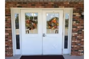 Double white doors with glass panels decorated with autumn-themed wreaths, set in a brick exterior wall with narrow vertical windows on each side and a dark doormat in front.