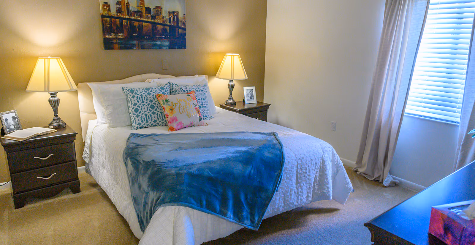 A neatly made bed with white bedding, decorative pillows, and a blue throw blanket in a bedroom. There are two bedside tables with lamps and framed photos on each side of the bed. A window with blinds and light curtains is on the right side of the room, and a piece of artwork depicting a city skyline hangs above the bed.