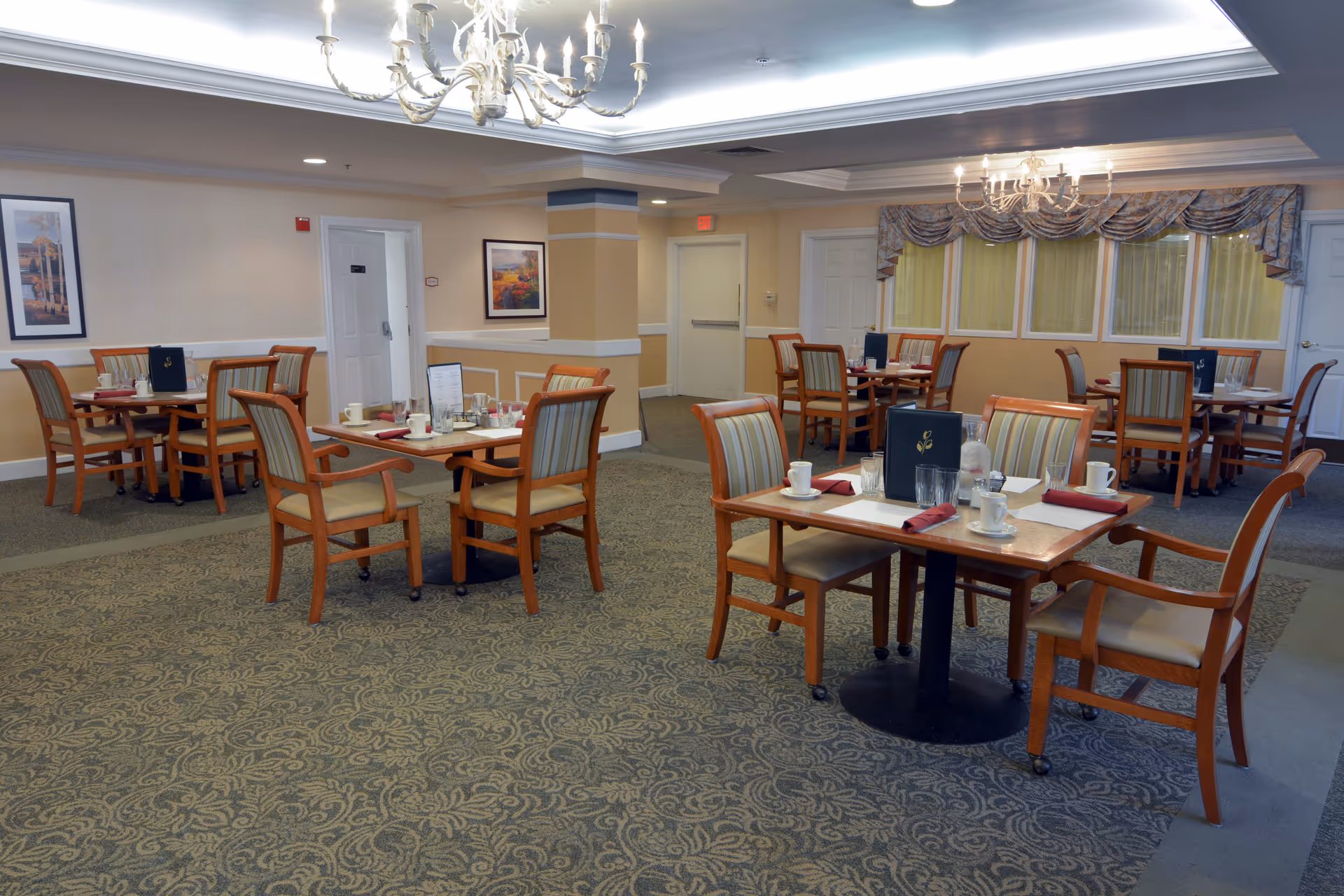 Bright dining room with multiple wooden tables and chairs set with cups, menus, and place settings under chandeliers.