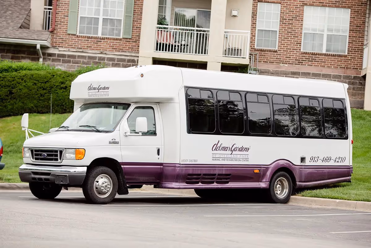A white and purple shuttle bus parked in front of a brick building with windows and balconies. The bus has the logo and contact information for Delmar Gardens of Overland Park Nursing and Rehabilitation Center on its side.