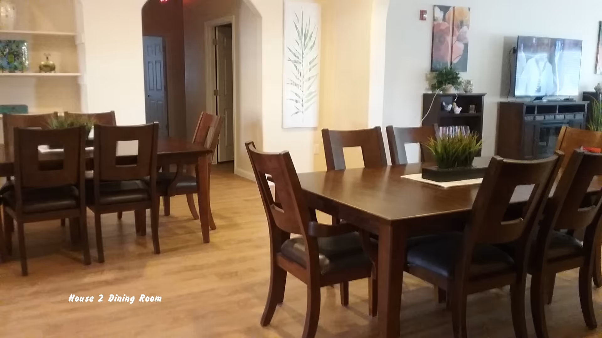 A dining room with two wooden dining tables surrounded by wooden chairs with dark cushions. The room has light-colored walls and wooden flooring. There is a plant centerpiece on one of the tables, wall art, and a television on a stand in the background.