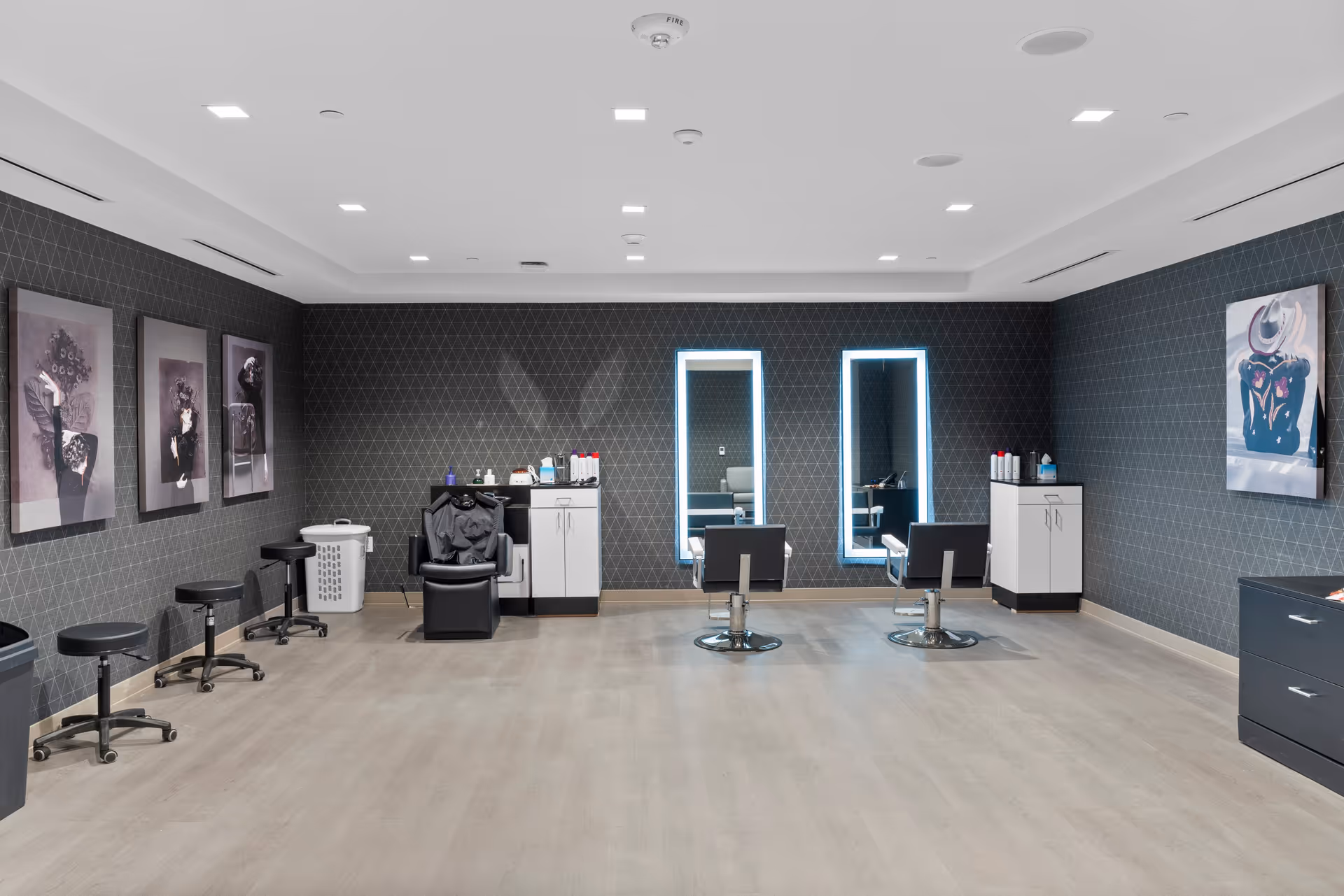Interior view of a modern hair salon with two styling chairs facing illuminated mirrors, a shampoo chair, stools along the left wall, and framed artwork on the walls. The room has light wood flooring and dark patterned wallpaper.