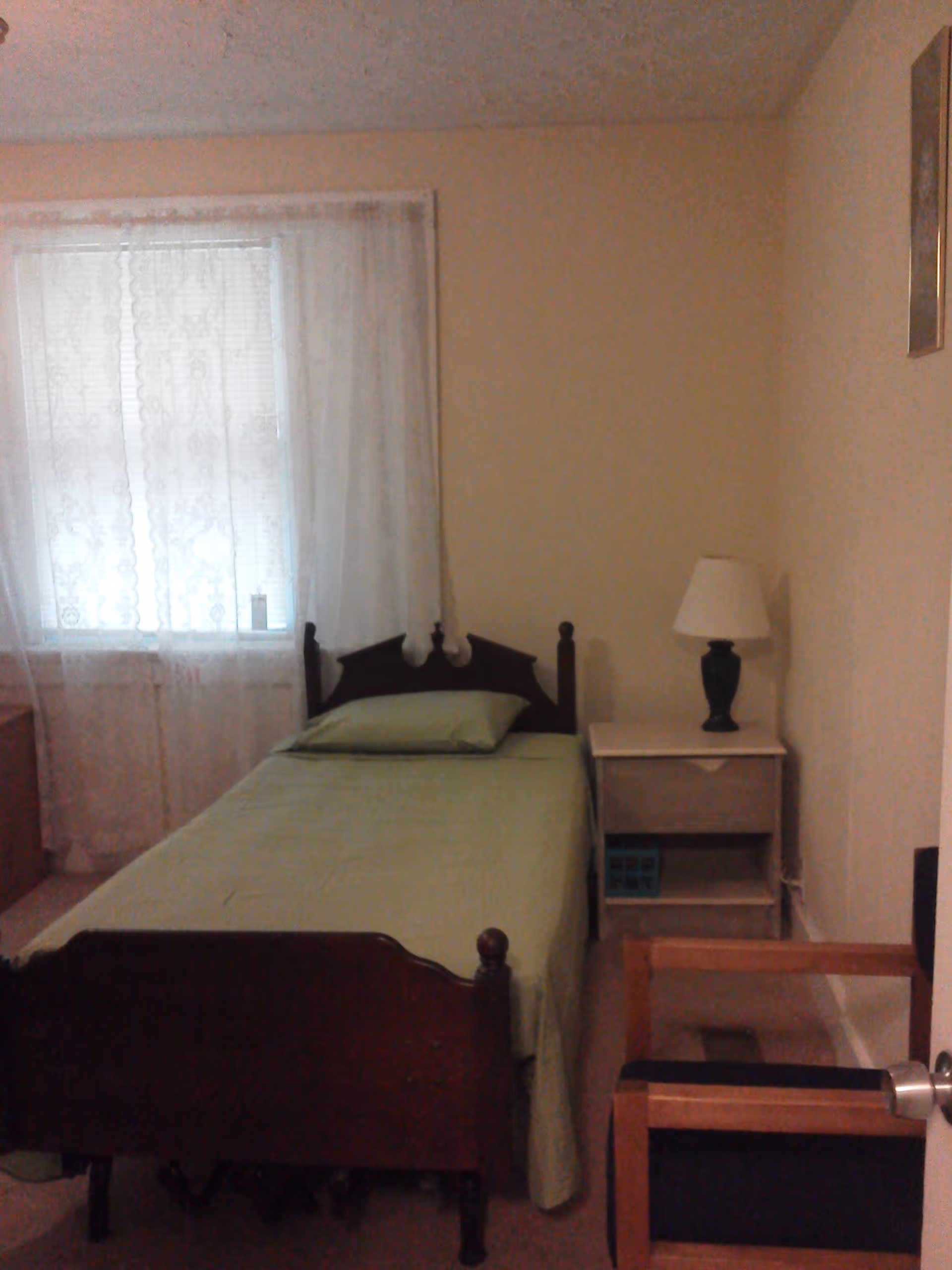 A small bedroom with a single wooden bed covered with a green bedspread and pillow. Next to the bed is a white nightstand with a dark green lamp on it. A window with white lace curtains is on the left wall, and a wooden chair with black cushions is partially visible in the foreground on the right side.