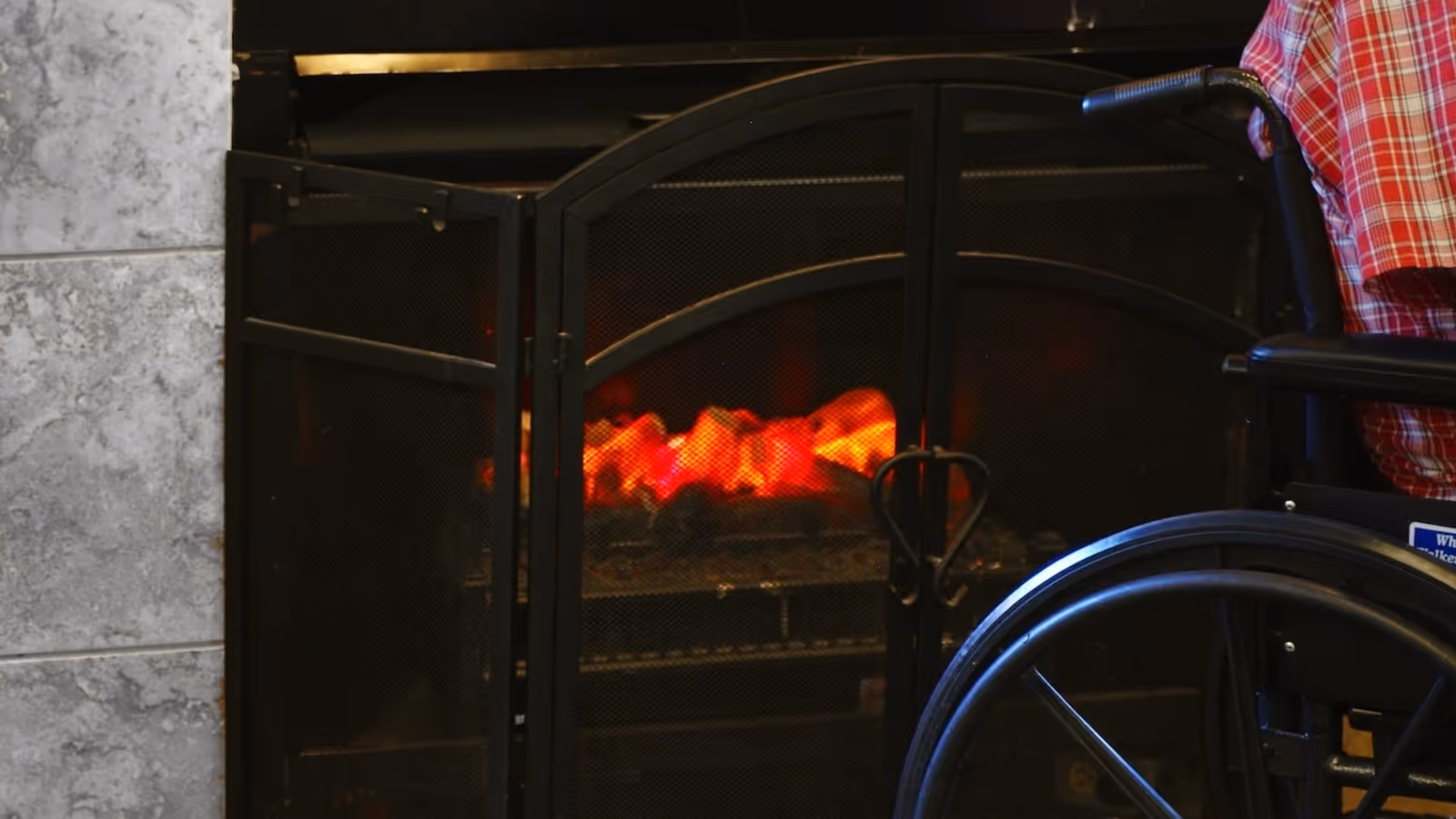 A close-up view of a fireplace with a black metal screen in front, showing glowing red and orange flames. To the right, part of a wheelchair with a red plaid blanket draped over the back is visible.