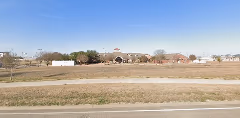 Wide view of a single-story building with a pitched roof and a small tower-like structure in the center, surrounded by leafless trees and a large grassy area in the foreground under a clear blue sky.