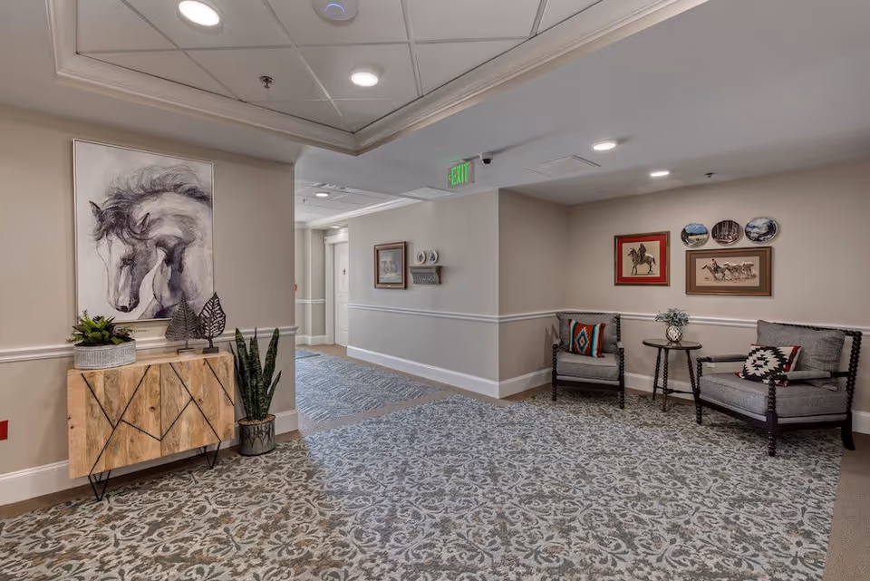 Carpeted senior living interior hallway with a wooden console and horse painting on the left and two upholstered chairs with a small table and wall art on the right.