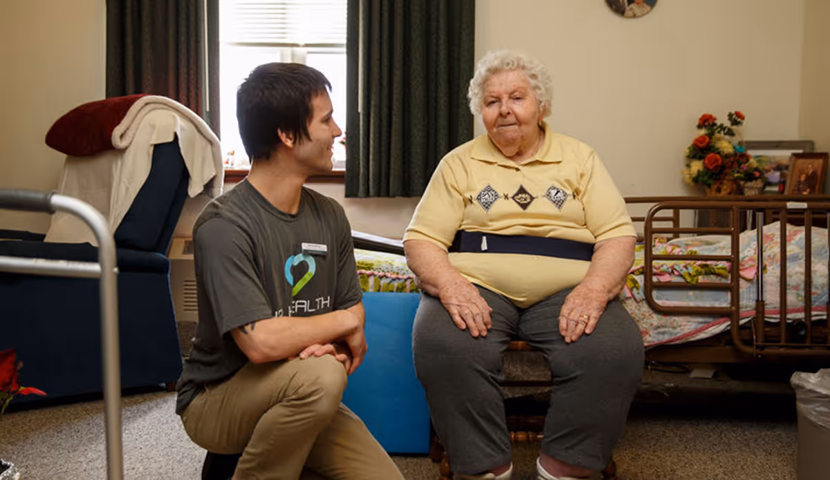 An elderly woman sitting on a chair in a room with a bed and floral decorations, while a younger man wearing a gray t-shirt with a heart logo kneels beside her, engaging in conversation.