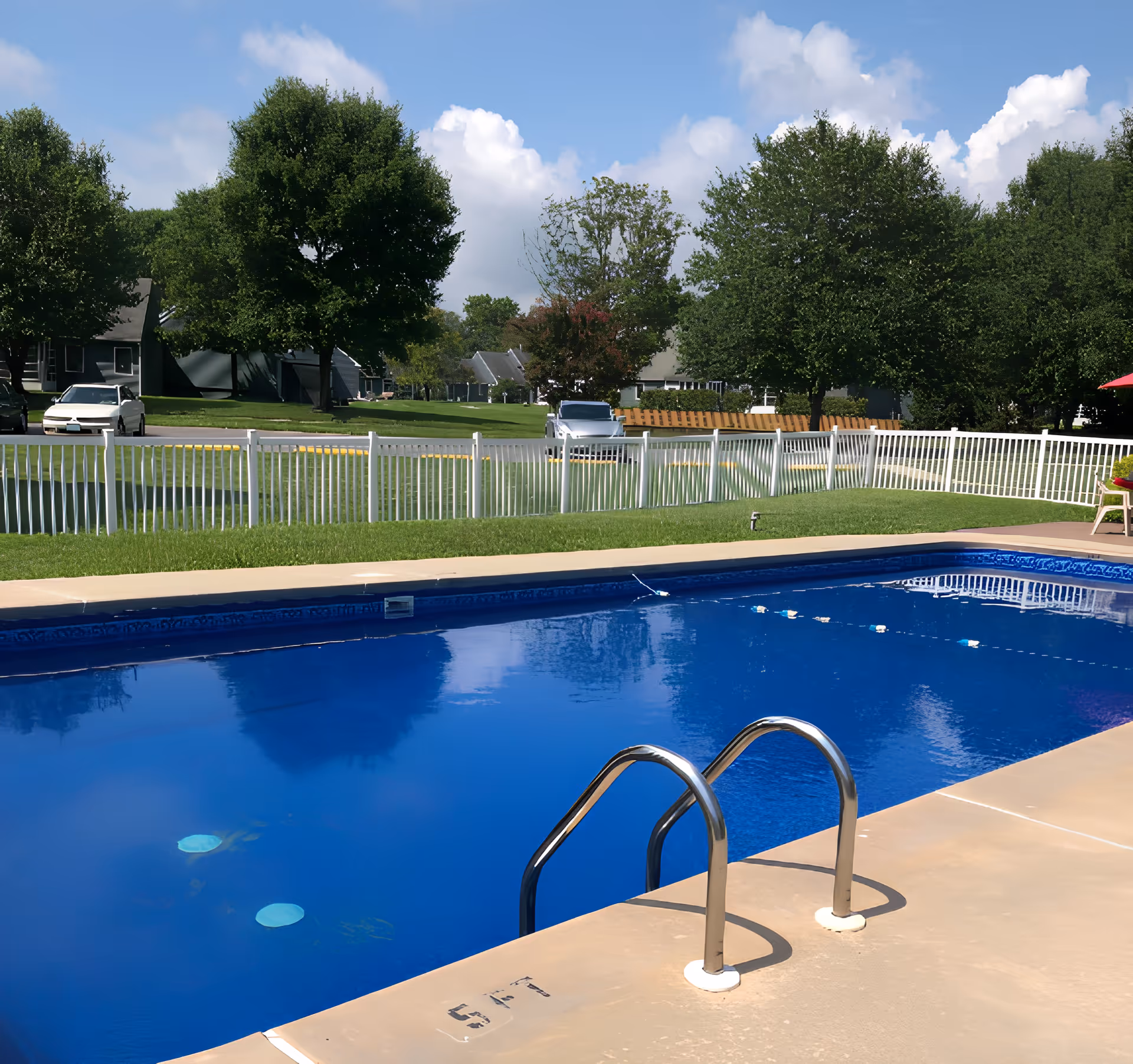 Outdoor community swimming pool with a metal ladder, white fence, lawn and houses with trees in the background.