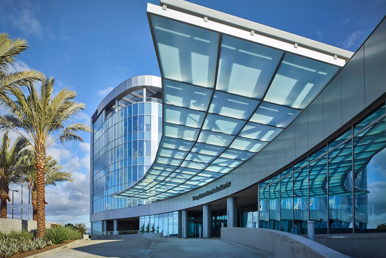 Curved glass-front modern medical building entrance with a large overhanging glass canopy and palm trees.