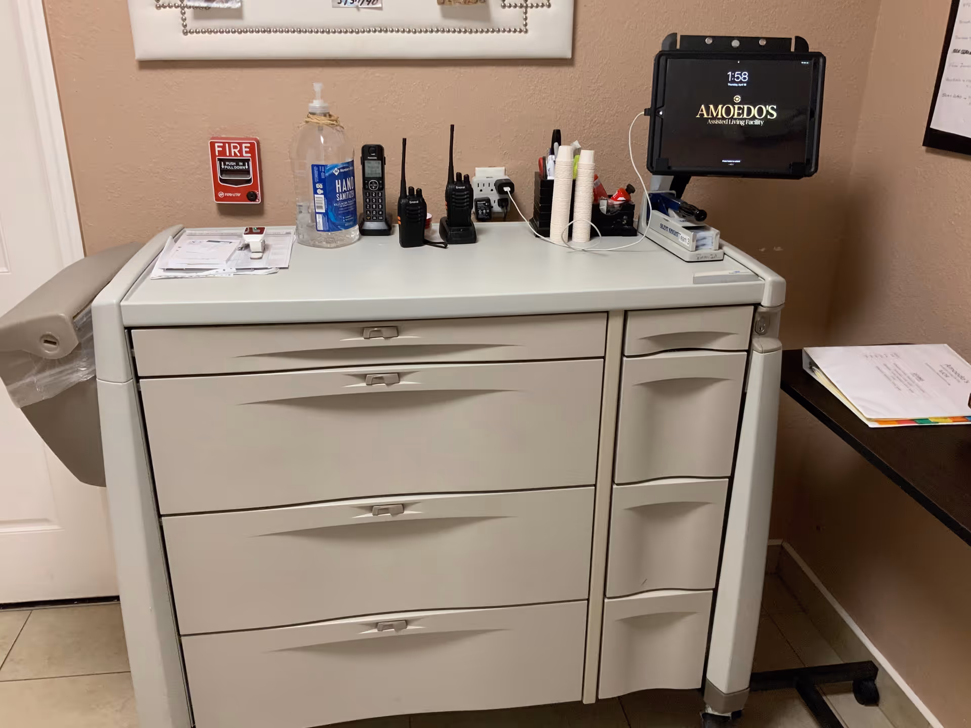 A beige medical supply cart with multiple drawers topped with walkie-talkies, a hand sanitizer bottle, papers and a tablet displaying 'Amoedo's' against a tan wall.