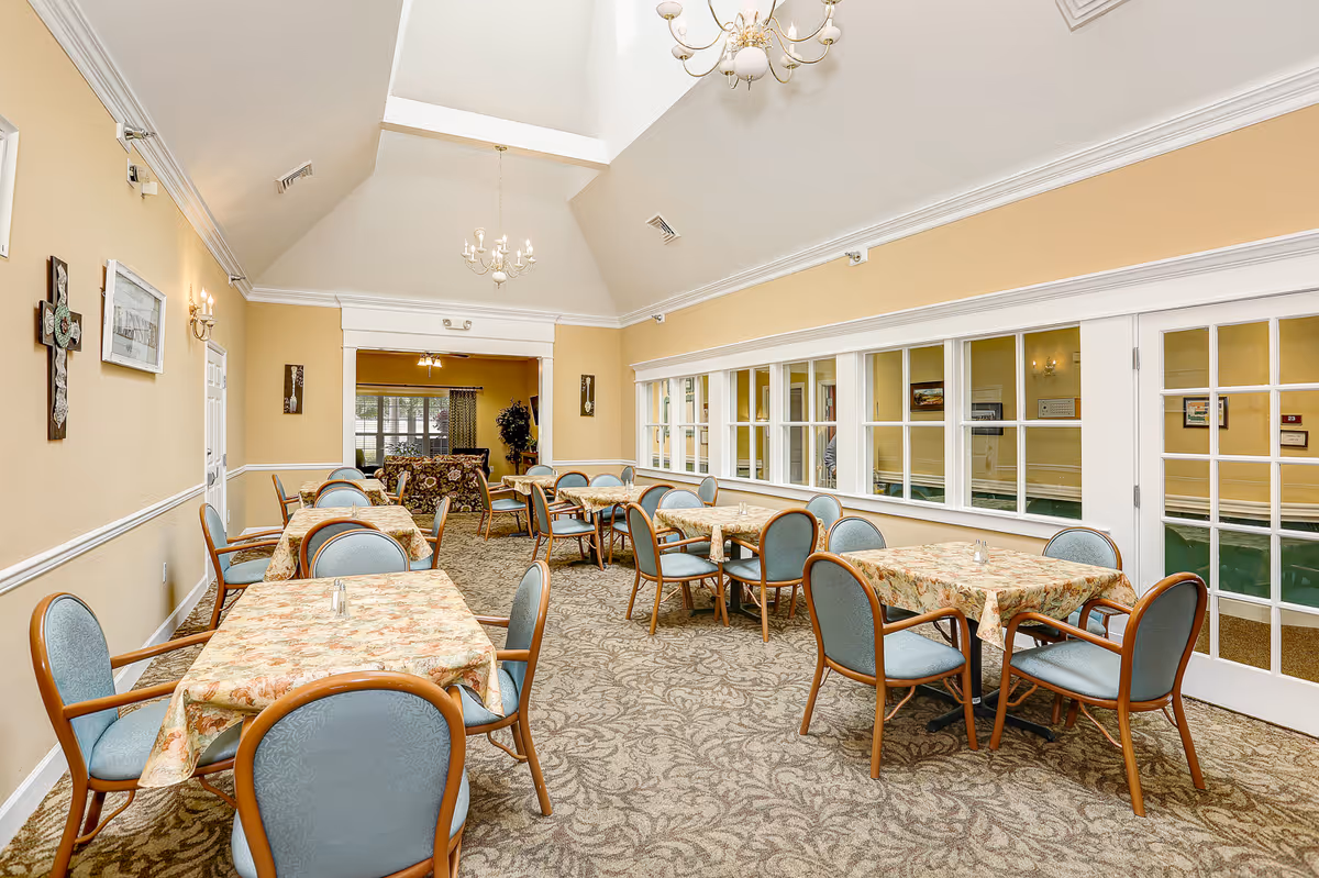A dining room with several tables covered in floral tablecloths and surrounded by blue cushioned chairs with wooden frames. The room has beige walls, a patterned carpet, and a high vaulted ceiling with chandeliers. There are windows and glass doors on one side, and a sitting area with a sofa is visible through an open doorway at the far end.