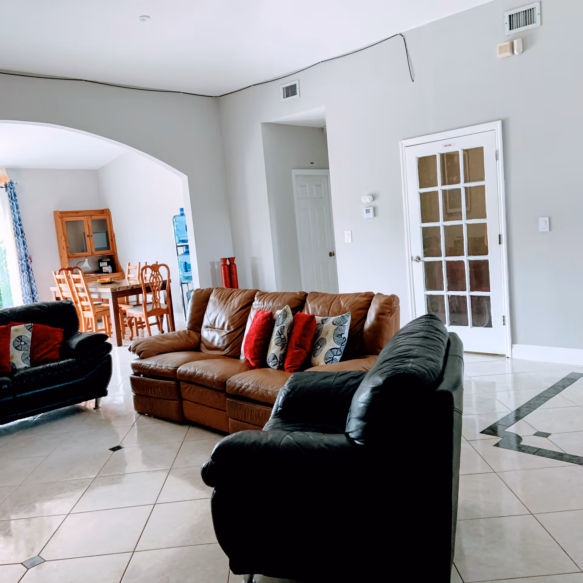 A living room with a brown leather sofa and a black leather armchair, both adorned with decorative pillows. The room has light-colored tiled flooring with a decorative pattern, light gray walls, and a white door with glass panels. In the background, there is a dining area with a wooden dining table and chairs, a wooden cabinet, and a water dispenser.