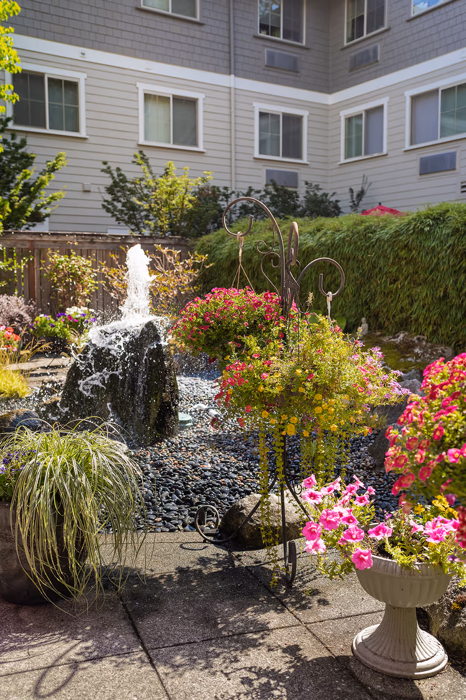 A bright outdoor garden area with colorful hanging flower baskets and potted plants surrounding a small water fountain. The background shows the exterior walls and windows of a multi-story building.