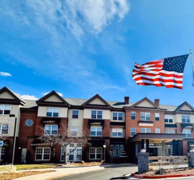 Exterior view of a multi-story senior living facility building with brick and beige siding under a blue sky with some clouds. An American flag is flying on a flagpole in front of the building.