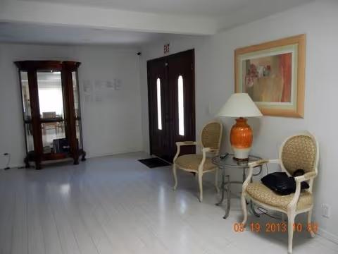 Small reception lobby with two upholstered chairs flanking a side table and lamp, framed art on the wall, a glass display cabinet, and double entry doors.