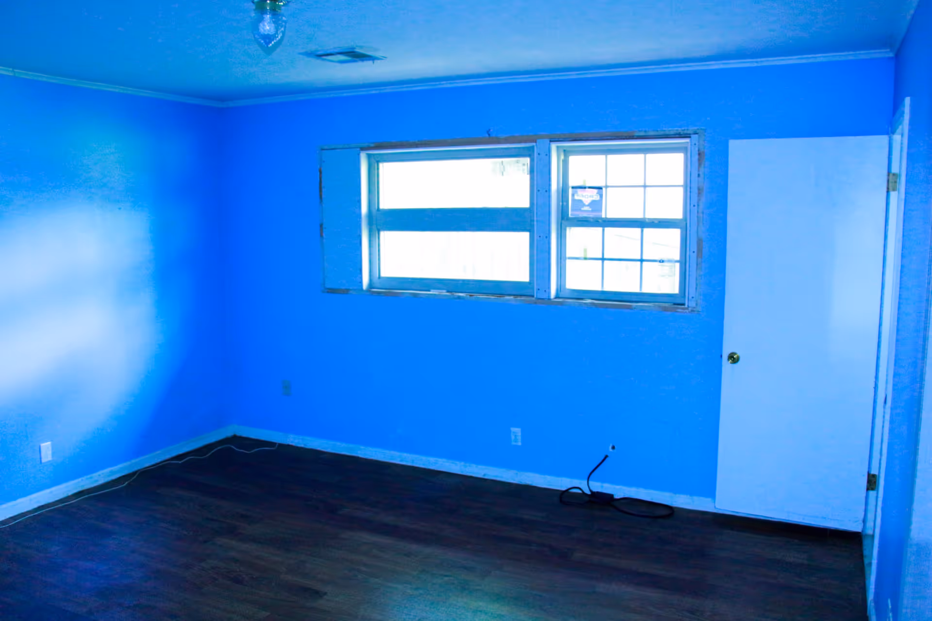 Empty room with bright blue walls, a window with two panes, a white door, and dark wood flooring. The room has a ceiling light fixture and some visible electrical outlets and cords on the floor.