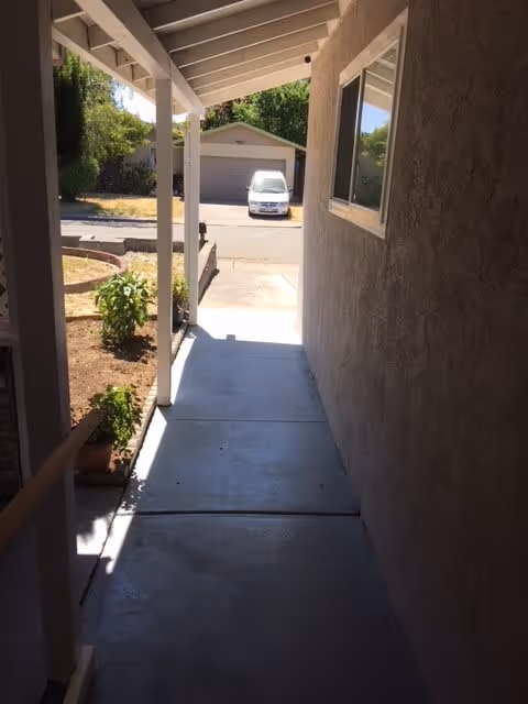 A covered walkway beside a building with a concrete path leading to a driveway. There are plants and a small garden area on the left side, and a white vehicle is parked in front of a garage across the street. The right side shows a textured wall with a window.