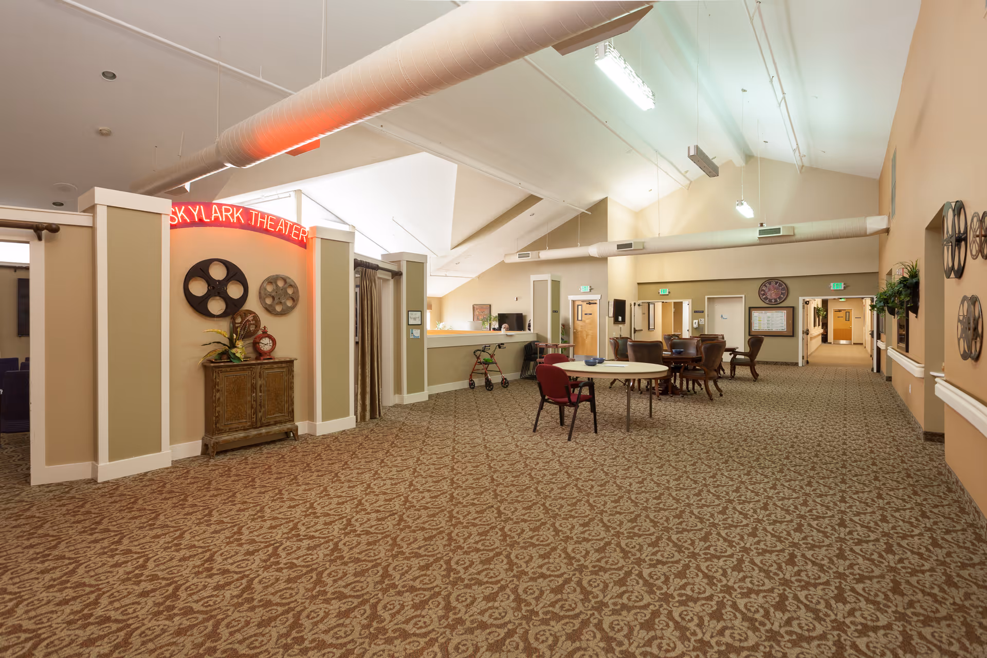 Interior view of a spacious common area in Skylark Assisted Living featuring patterned carpet flooring, a round table with chairs, a small wooden cabinet with decorative items, and a neon sign reading 'Skylark Theater'. The area has high ceilings with exposed ductwork and multiple doorways leading to other rooms.