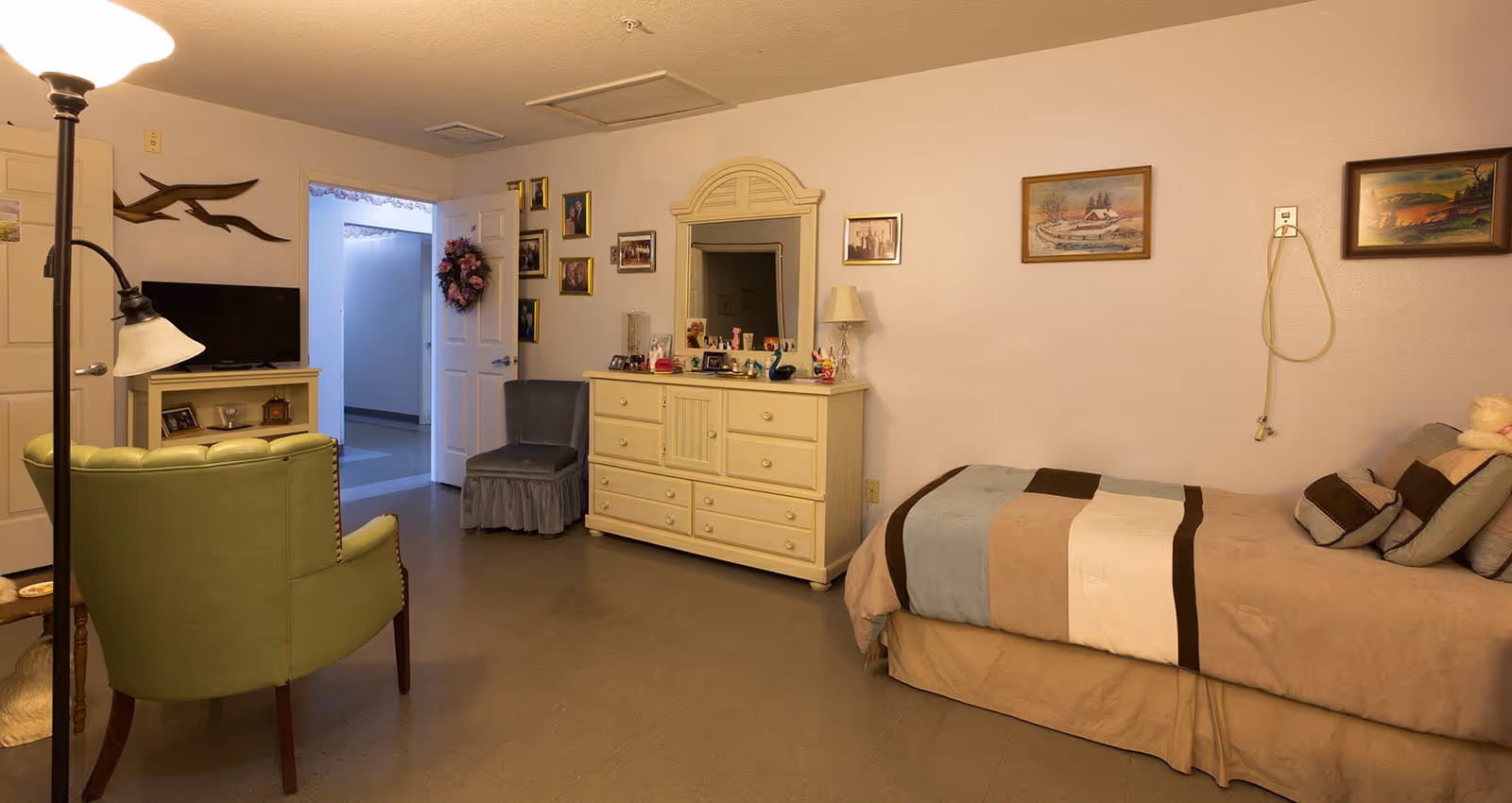 A cozy assisted living bedroom with a single bed covered in a patchwork quilt, a beige dresser with a mirror, a small lamp, and various framed photos and decorations on the walls. There is a green armchair and a small TV on a stand near the door, which is open to a hallway. The room has a warm, inviting atmosphere.