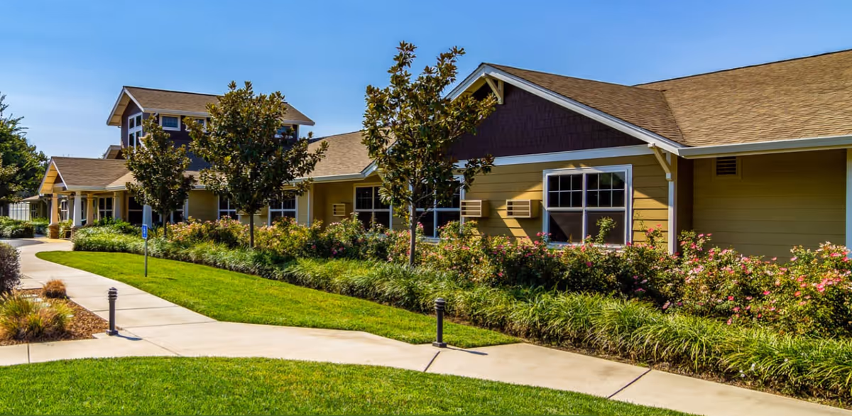 Single-story yellow building with a landscaped lawn, flowering shrubs, and a curved sidewalk under a clear blue sky.