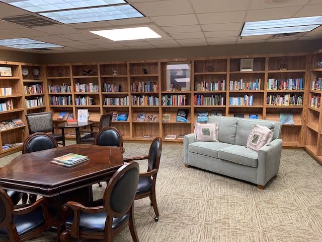 A cozy library room with wooden bookshelves lining the walls filled with books. There is a light gray loveseat with two decorative pillows on the right side and a wooden table with four chairs in the center. The room has a beige carpet and a drop ceiling with fluorescent lighting.